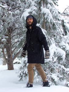 Ein junger bärtiger Mann stapft durch den hohen Schnee in einem Wald. Es Schneit. Der Mann trägt Winterkleidung, Handschuhe und Barfuß Schneestiefel der Marke Xero Shoes.