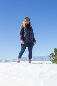 Eine junge blonde Frau steht bei Sonnenschein im Schnee. Im Hintergrund sieht man Berge. Sie lächelt und trägt Winterkleidung, inklusive der Schneestiefel von Xero Shoes.