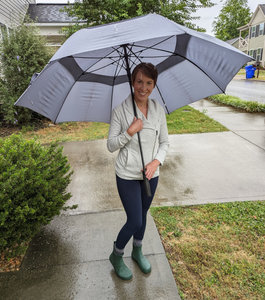 Eine Frau steht auf einem asphaltiertem Fußweg im Regen. Sie trägt einen großen grauen Regenschirm, eine beige Jacke, blaue Hose und grüne Barfußschuhe Gummistiefel Modell Xero Shoes Gracie Hunter. Sie lächelt. Im Hintergrund sieht man Büsche, Rasen und ein helles Haus mit dunklem Dach.