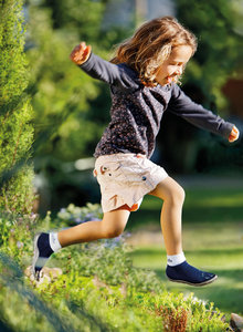 Ein Kind in kurzer beiger Hose und blauem Oberteil mit Pünktchen drauf springt über eine niefrige Hecke. Es trägt blaue Barfuß-Kinderschuhe vom Modell Leguanito blau.