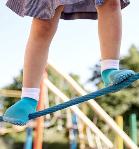 Ein Kind balanciert auf einem Spielplatz über ein Seil. Es trägt ein Röckchen und Barfuß-Kinderschuhe vom Modell Leguanito türkis.