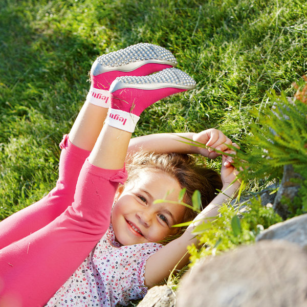 Ein Mädchen liegt im Gras und lächelt den Betrachter an. Es trägt eine pinke Hose, ein gepunktetes Oberteil und pinke Kinder Barfußschuhe. Es reckt die Füße Richtung Himmel und verschränkt die Arme überm Kopf.