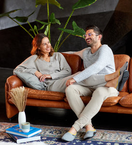Ein Pärchen sitzt lachend auf einem Ledersofa. Sie tragen helle Klamotten, vielleicht Schlafanzüge und graue Barfuß-Hausschuhe vom modell Leguano Acasa.