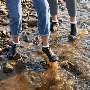 2 Personen laufen in Barfuß-Sockenschuhe an einem steinigen Ufer durch das flache Wasser. Sie haben die Hosen hochgekrempelt und tragen Barfuß-Minimalschuhe des Modell Leguano Classic schwarz, weißer Bund.