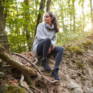 Eine junge Frau sitzt in einem herbstlichen Wald an einer Böschung. Die Sonne scheint und der Boden ist laubbedeckt. Sie trägt einen grauen Pullover, eine schwarze Hose und schwarze Winter-Barfußstiefel, Modell Leguano Kosmo schwarz. 